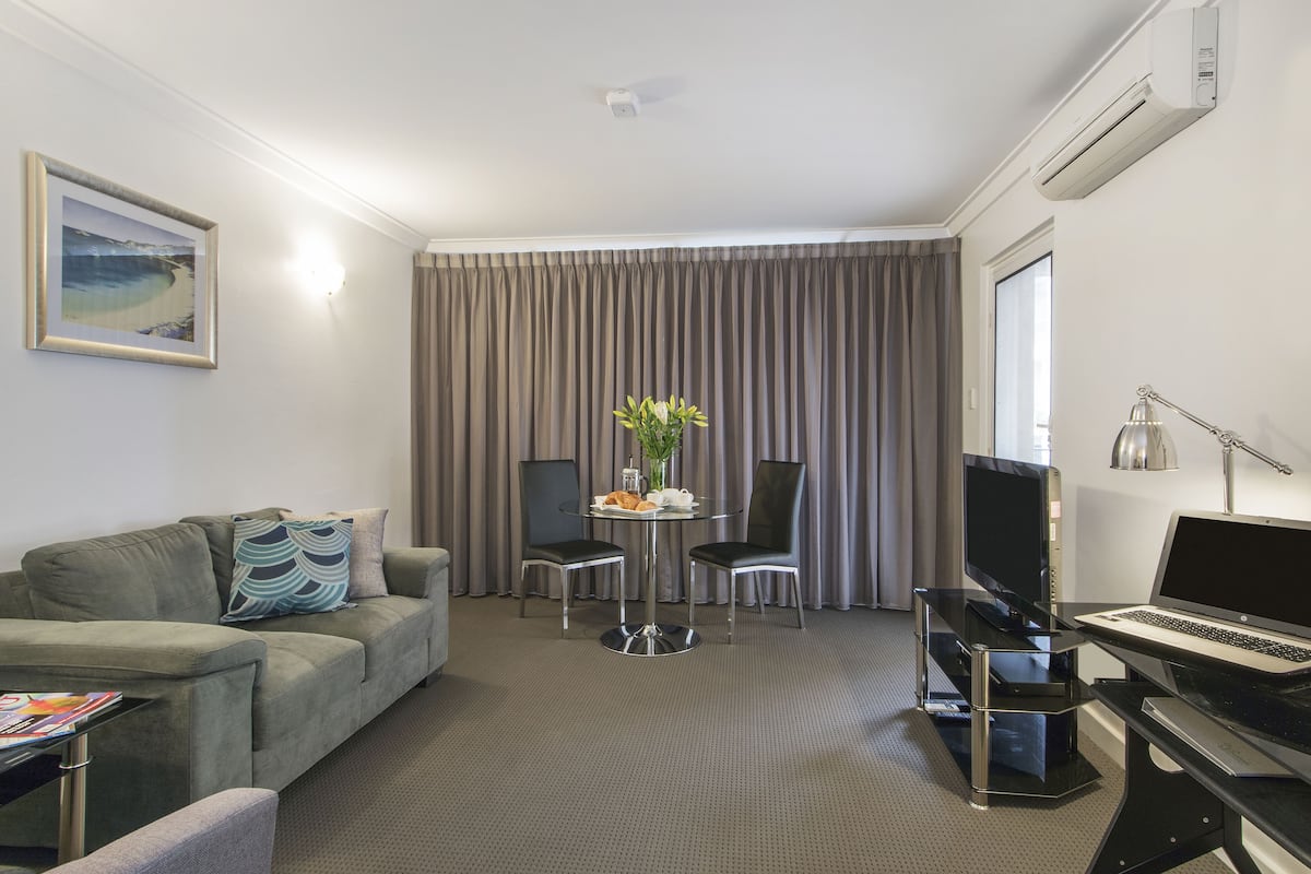 A hotel room with a grey sofa, a small round table with a vase and flowers, a TV, a laptop, and a desk lamp.
