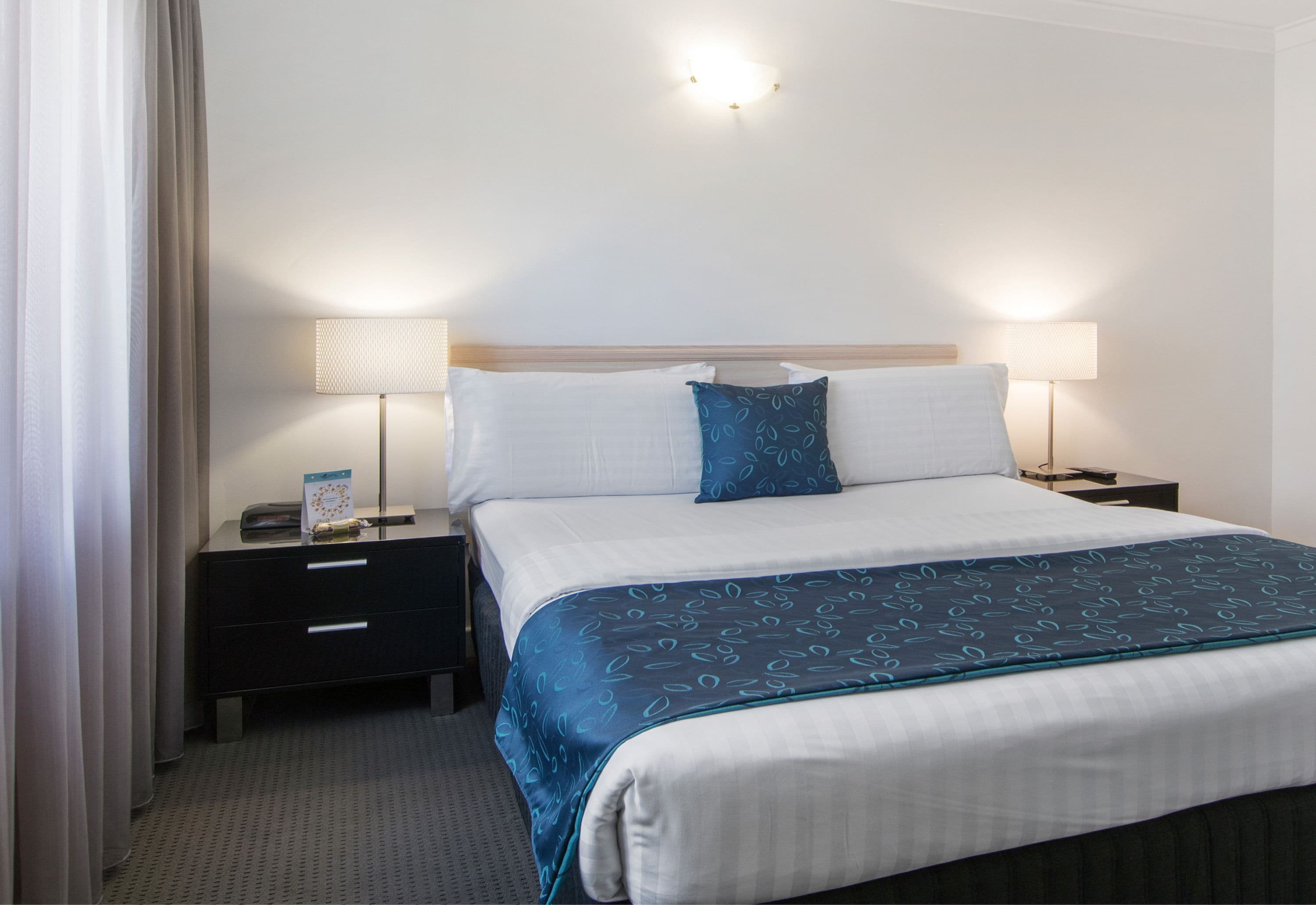 A hotel room with a neatly made bed, two bedside tables with lamps, a dark-colored dresser, and a light-colored headboard.