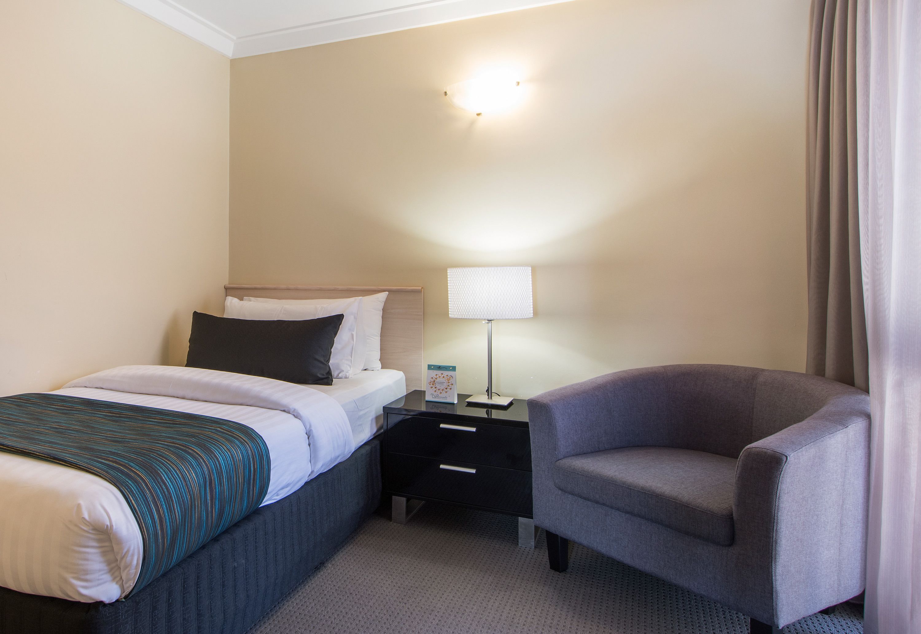 Une chambre d’hôtel avec un lit, une table de chevet avec une lampe, une chaise et une fenêtre avec des rideaux.