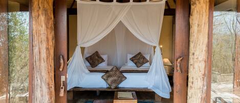Una cama con estructura de madera y dosel, dos almohadas y una pequeña mesa de madera en una habitación con vista a árboles.