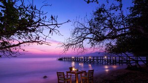 Une scène de plage paisible, avec une table et des chaises en bois sous un ciel crépusculaire, donnant sur des eaux calmes et une jetée lointaine.