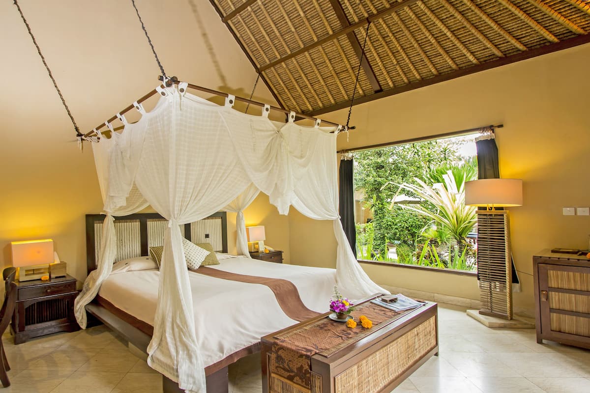 Une chambre à coucher avec un lit à baldaquin, une table de chevet avec une lampe, une commode et une vue sur la verdure à l’extérieur.