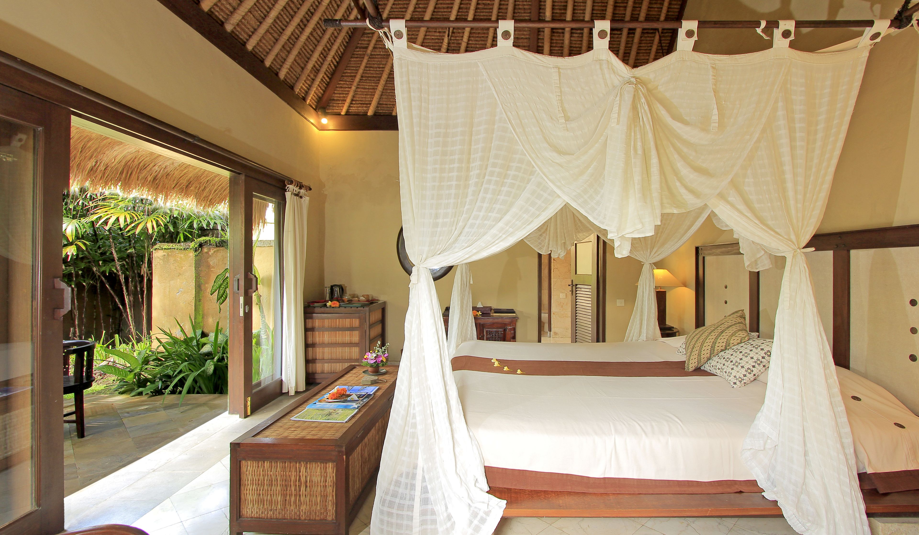 Habitación con una cama bajo un dosel, un banco de madera y vista a un jardín por una puerta de cristal.