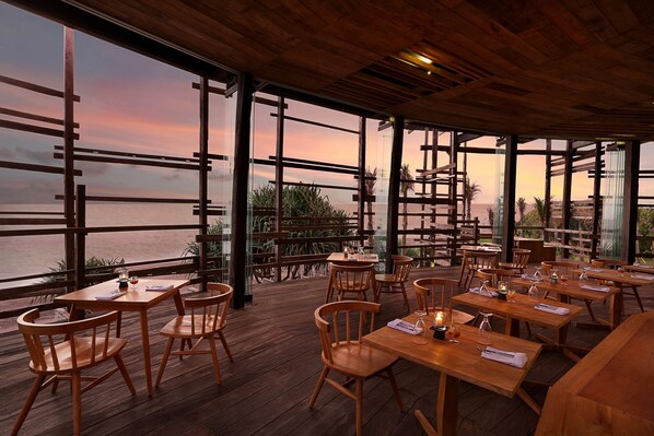 Ein Restaurant im Freien mit Holztischen und -stühlen, großen Fenstern und Meerblick.