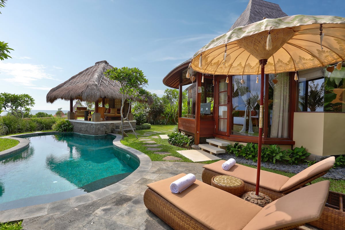 Un espace aménagé au bord de la piscine, avec des chaises longues, une structure à toit de chaume et un bâtiment moderne surmonté d’un grand parasol.