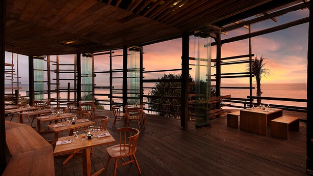 An outdoor dining area with wooden tables and chairs, large windows offering a view of the sunset over the ocean, and a wooden ceiling structure. - WakaGangga (Tabanan)