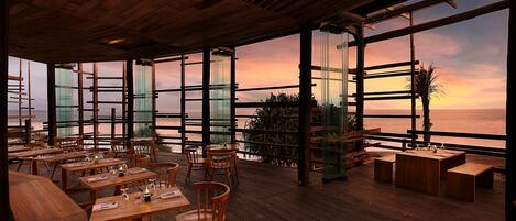 An outdoor dining area with wooden tables and chairs, large windows offering a view of the sunset over the ocean, and a wooden ceiling structure.