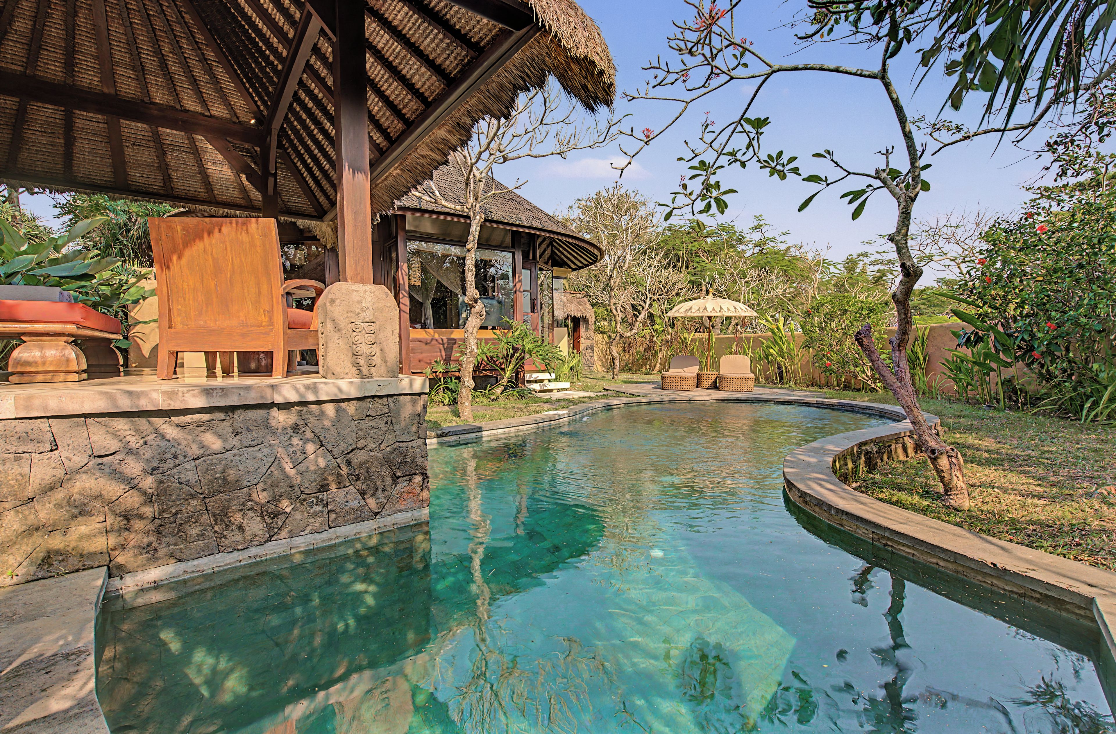 A clear pool with a stone wall, wooden furniture, and a thatched-roof structure.
