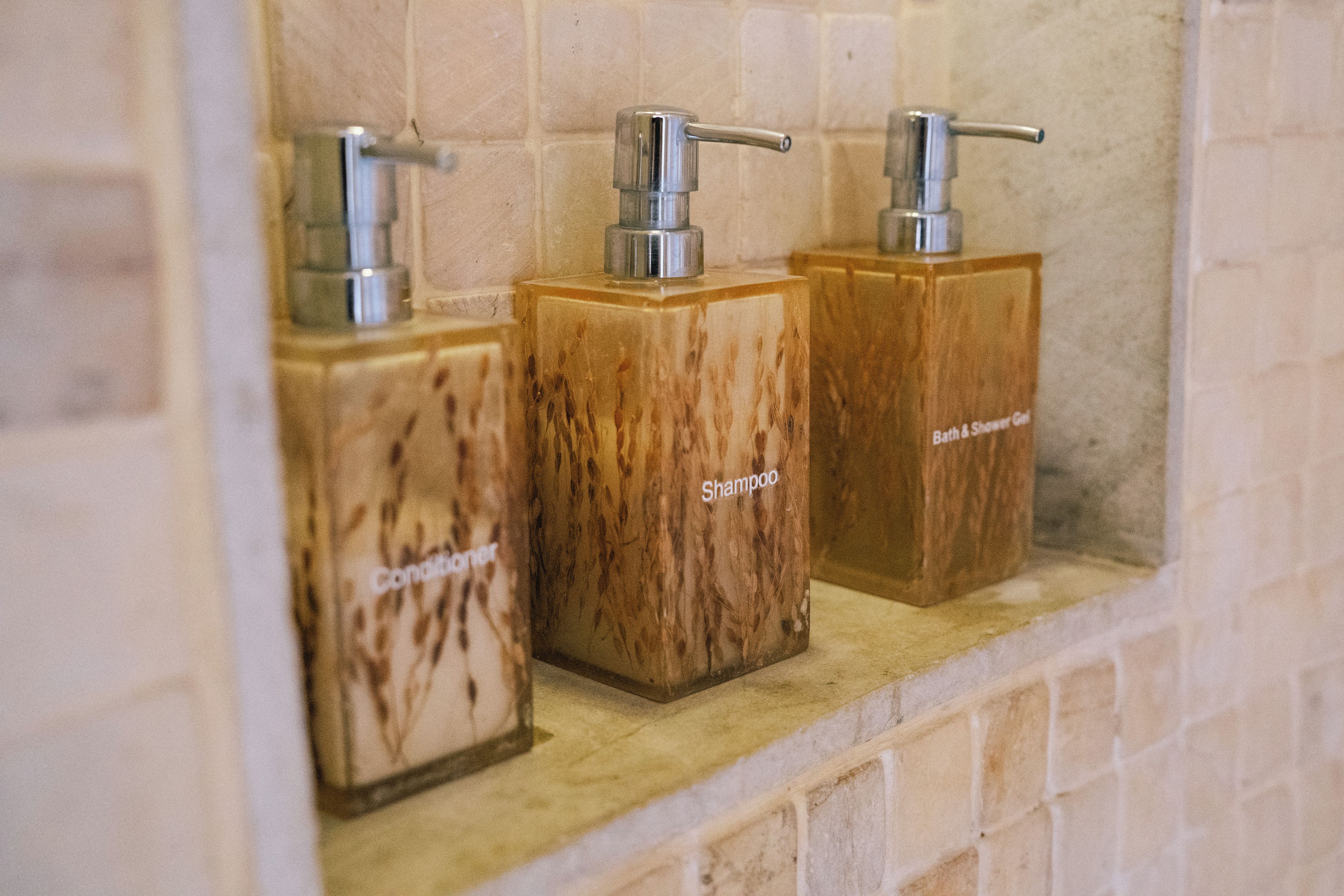 Tres dispensadores de madera con las etiquetas 'Champú' y 'Gel de Baño y Ducha' en una pared de azulejos.