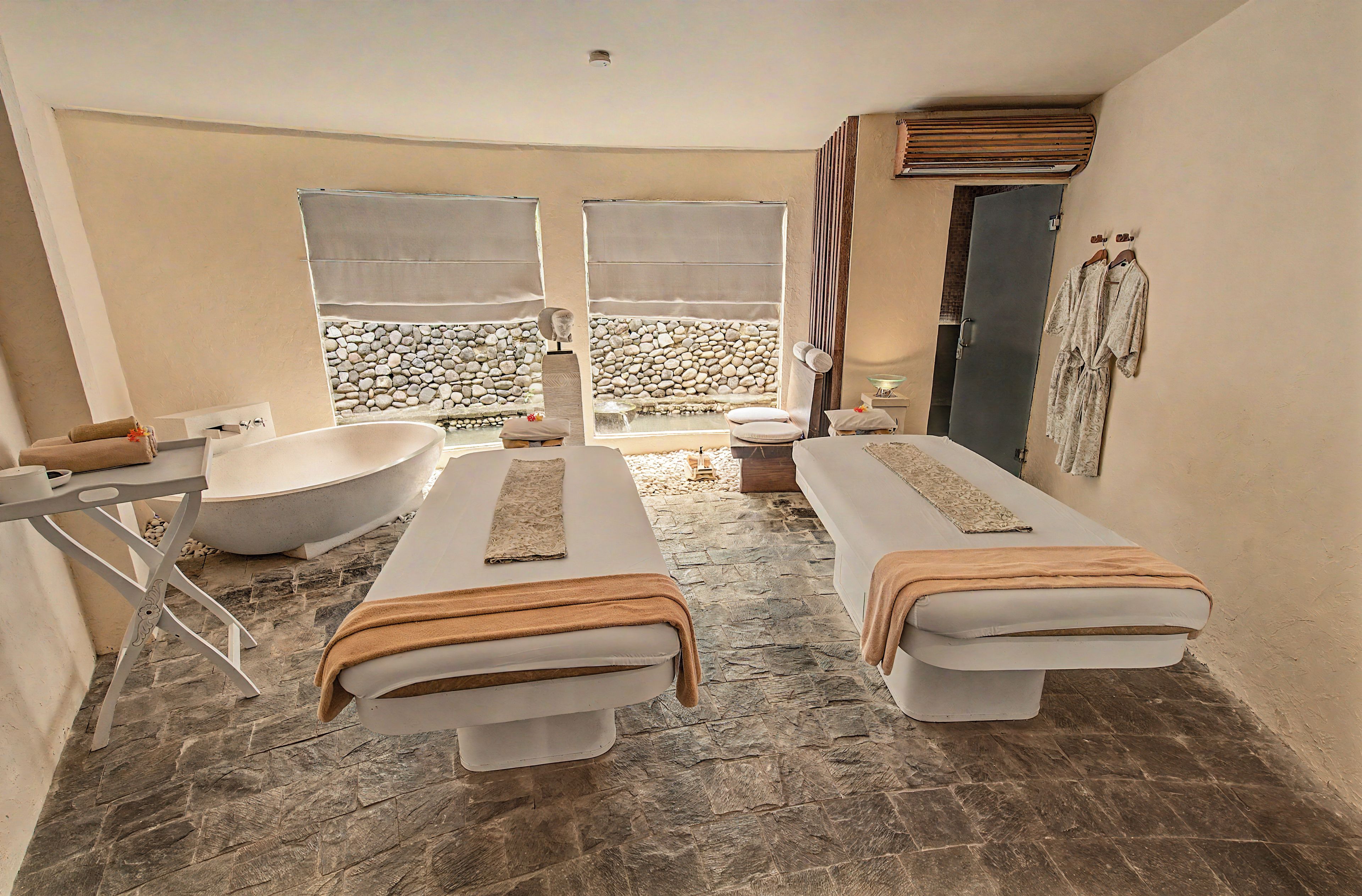 A spa room with two massage tables, a bathtub, and a stone wall backdrop.