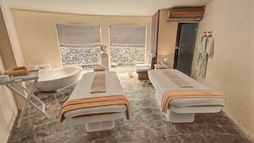 A spa room with two massage tables, a bathtub, and a stone wall backdrop.