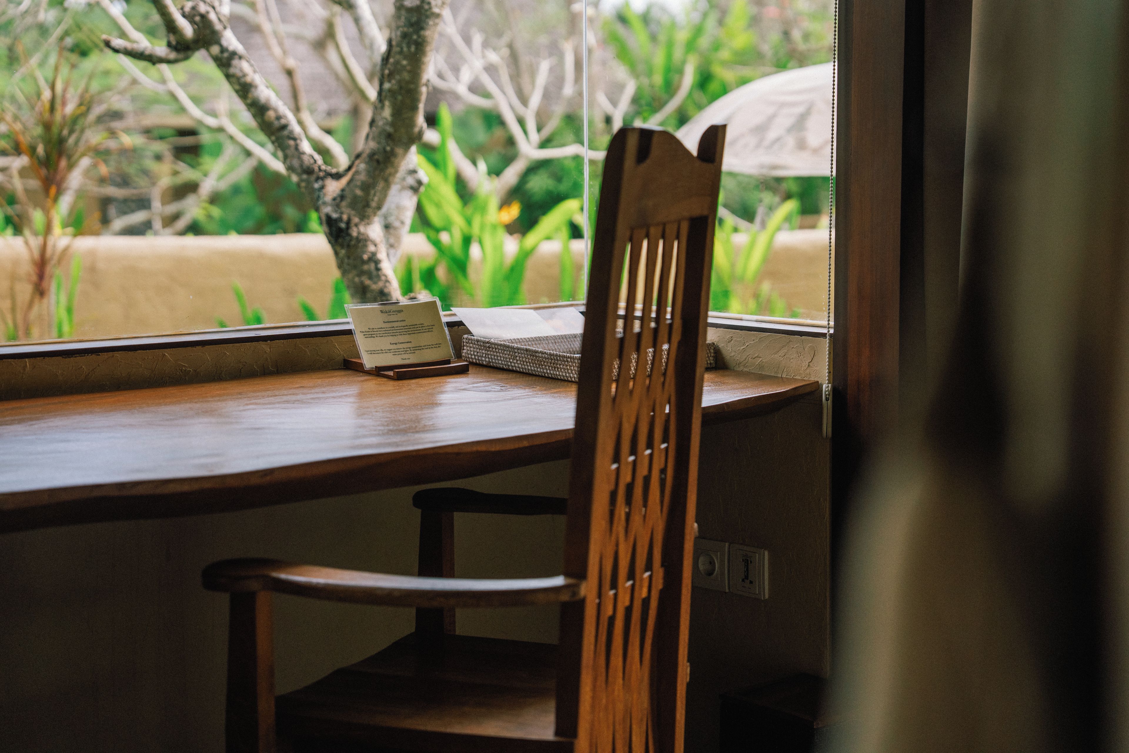 Una mesa de madera con una silla, un letrero pequeño y una ventana que da a zonas verdes.