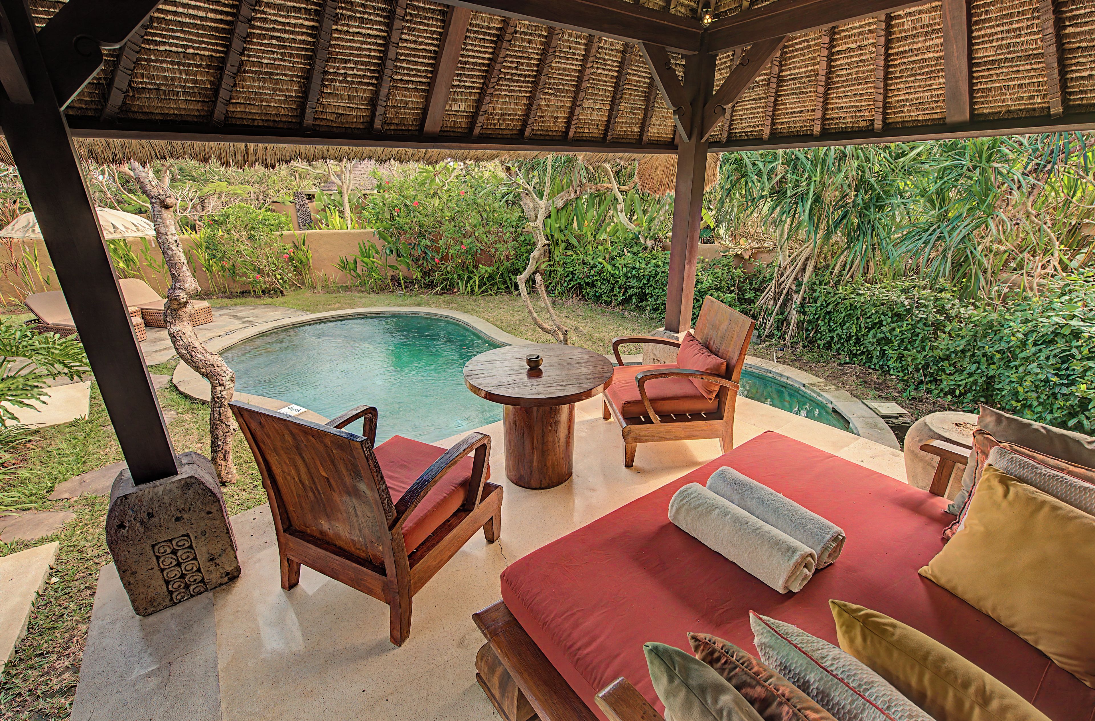 A covered outdoor area with a pool, wooden furniture, and lush greenery.