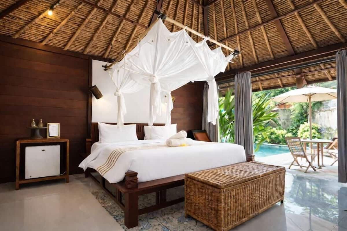 A bedroom with a canopy bed, a bedside table with a lamp, a small bedside cabinet, and a wicker ottoman.