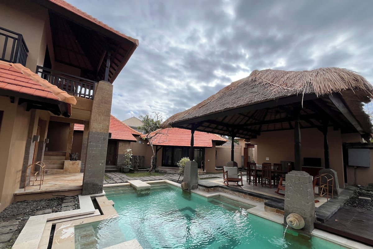 A pool area with a thatched roof structure, stone steps leading to a pool, and a seating area with chairs and tables.