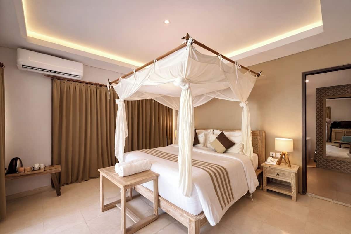 A four-poster canopy bed with a bedside table and lamp, a mirror, and a view of the adjacent room.