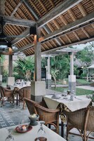 An outdoor dining area with wicker chairs and tables, a thatched roof, and a garden view.