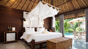 A bedroom with a canopy bed, a small bedside table, a nightstand, a wicker ottoman, and a view of an outdoor pool area. - Visakha Sanur by Puri Signatures (Denpasar)