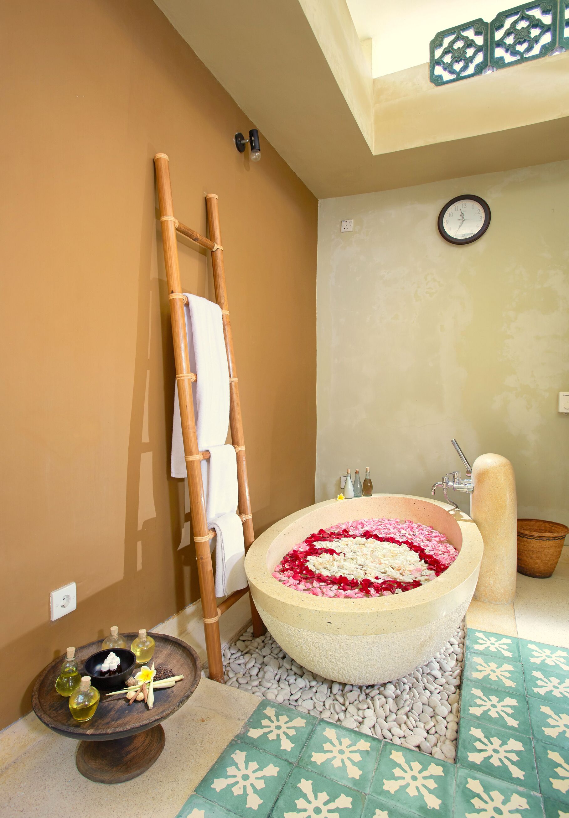 A bathroom with a large bathtub filled with rose petals, a wooden ladder, a clock on the wall, and a small table with bottles and a bowl.