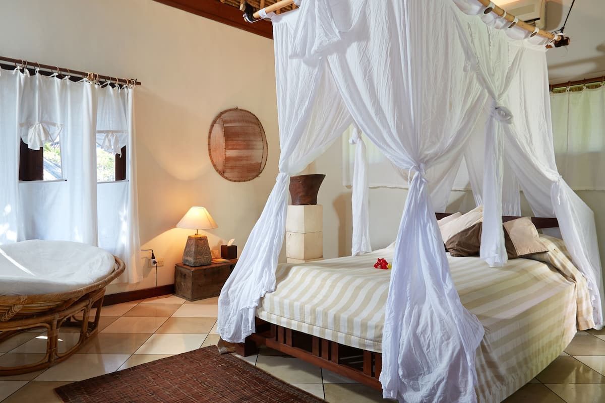 A room with a bed under a canopy, a wicker chair, a lamp, and a textured rug.
