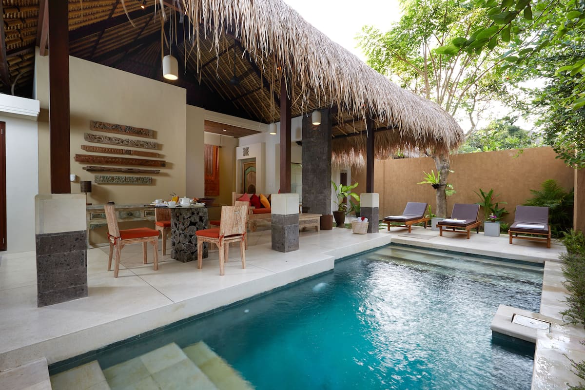 A pool area with lounge chairs, a table, and chairs under a thatched roof.