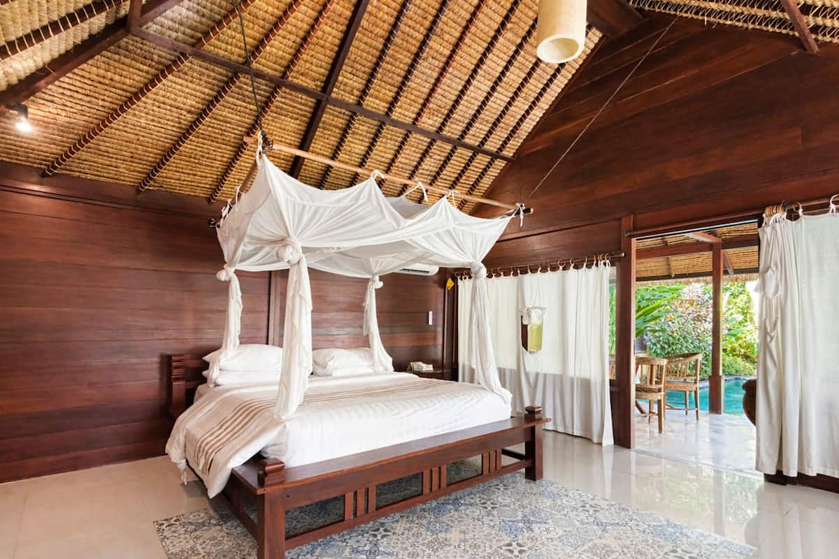 A bedroom with a four-poster bed, wooden walls, a tiled floor, and a thatched ceiling.