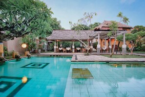 A swimming pool with a tiled deck, a covered seating area with chairs, and a building with a thatched roof. - Visakha Sanur by Puri Signatures (Denpasar)