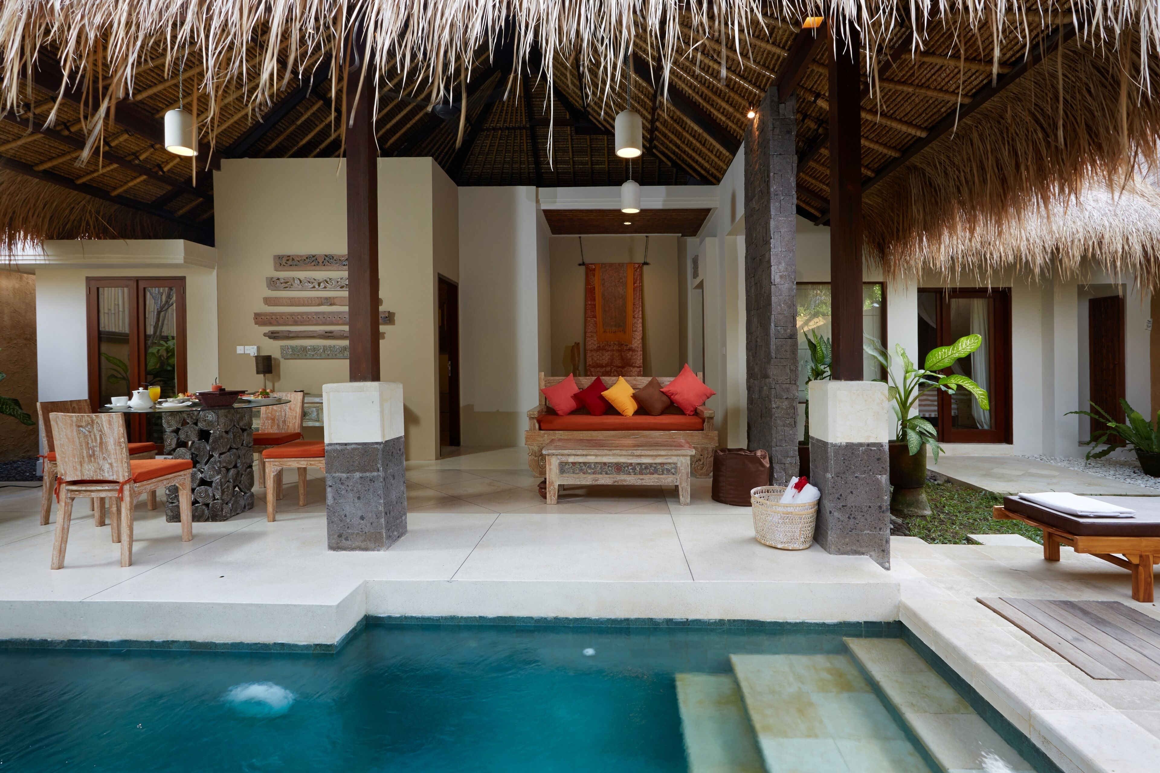 A poolside area with a thatched roof, wooden furniture, and a stone wall.