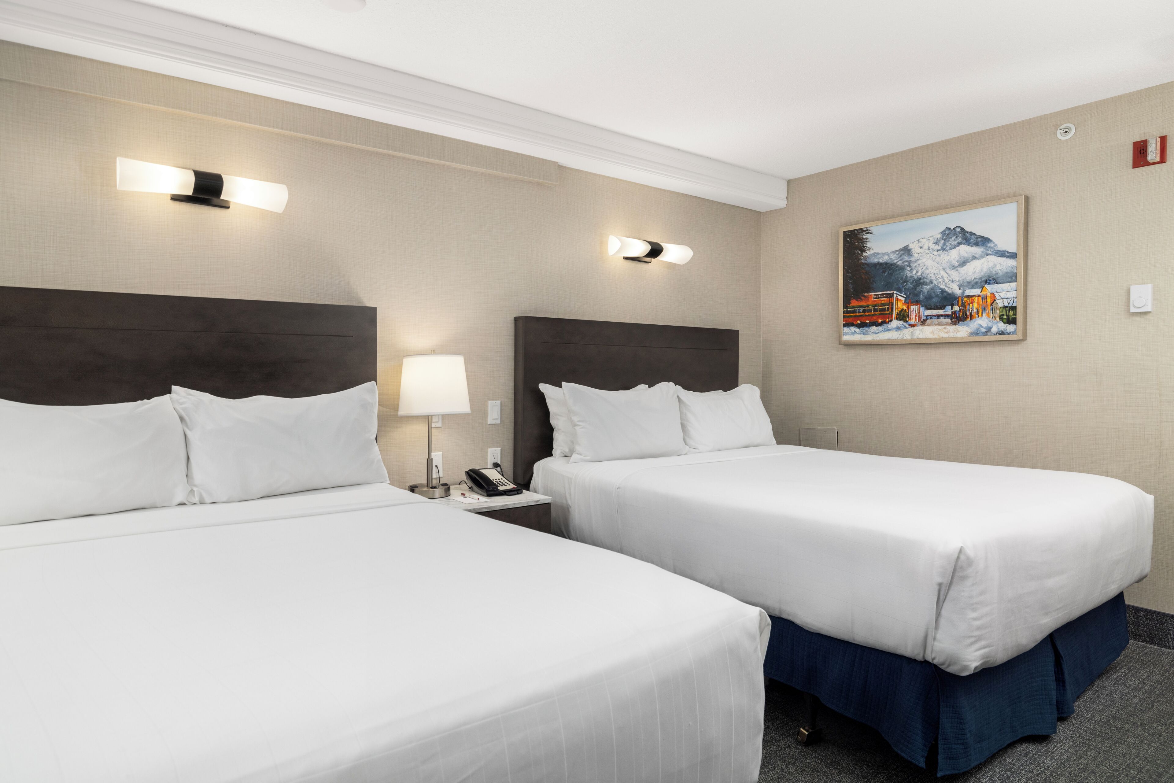 A hotel room with two beds, a nightstand, a lamp, a painting, and a thermostat on the wall.