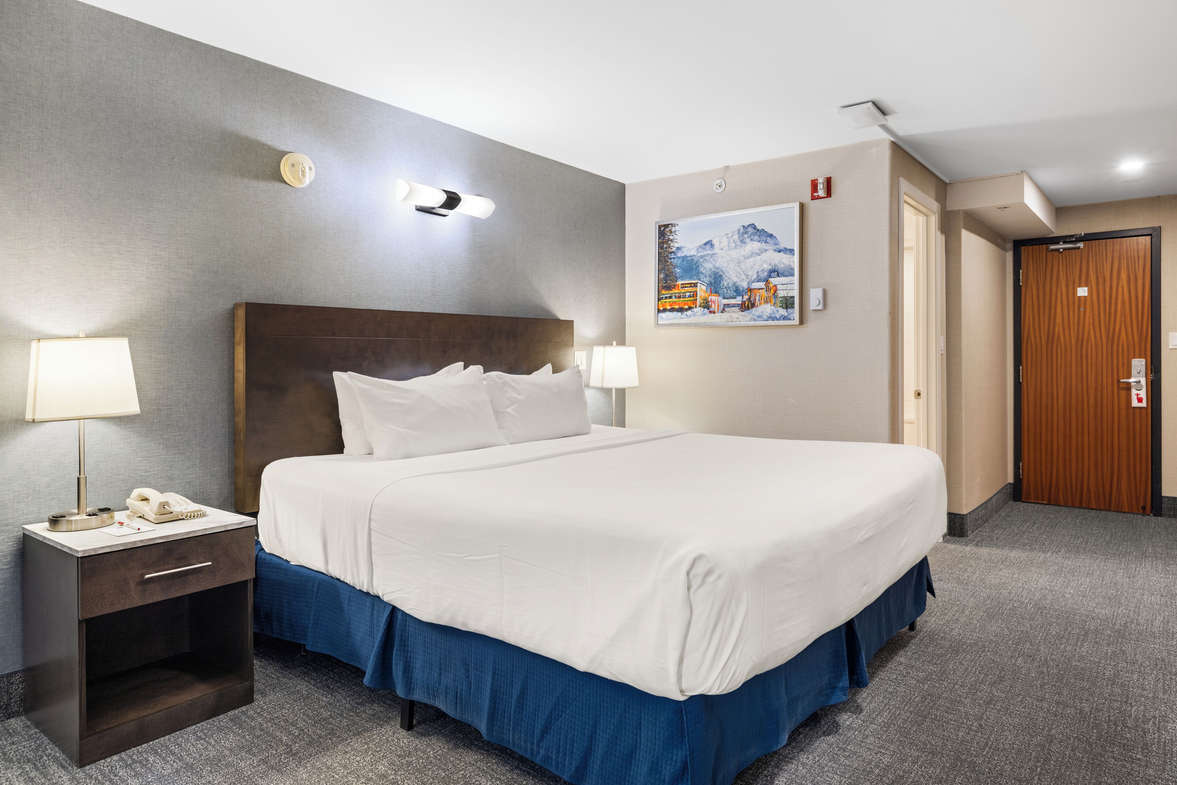 A hotel room with a bed, bedside table, lamp, and a painting on the wall.