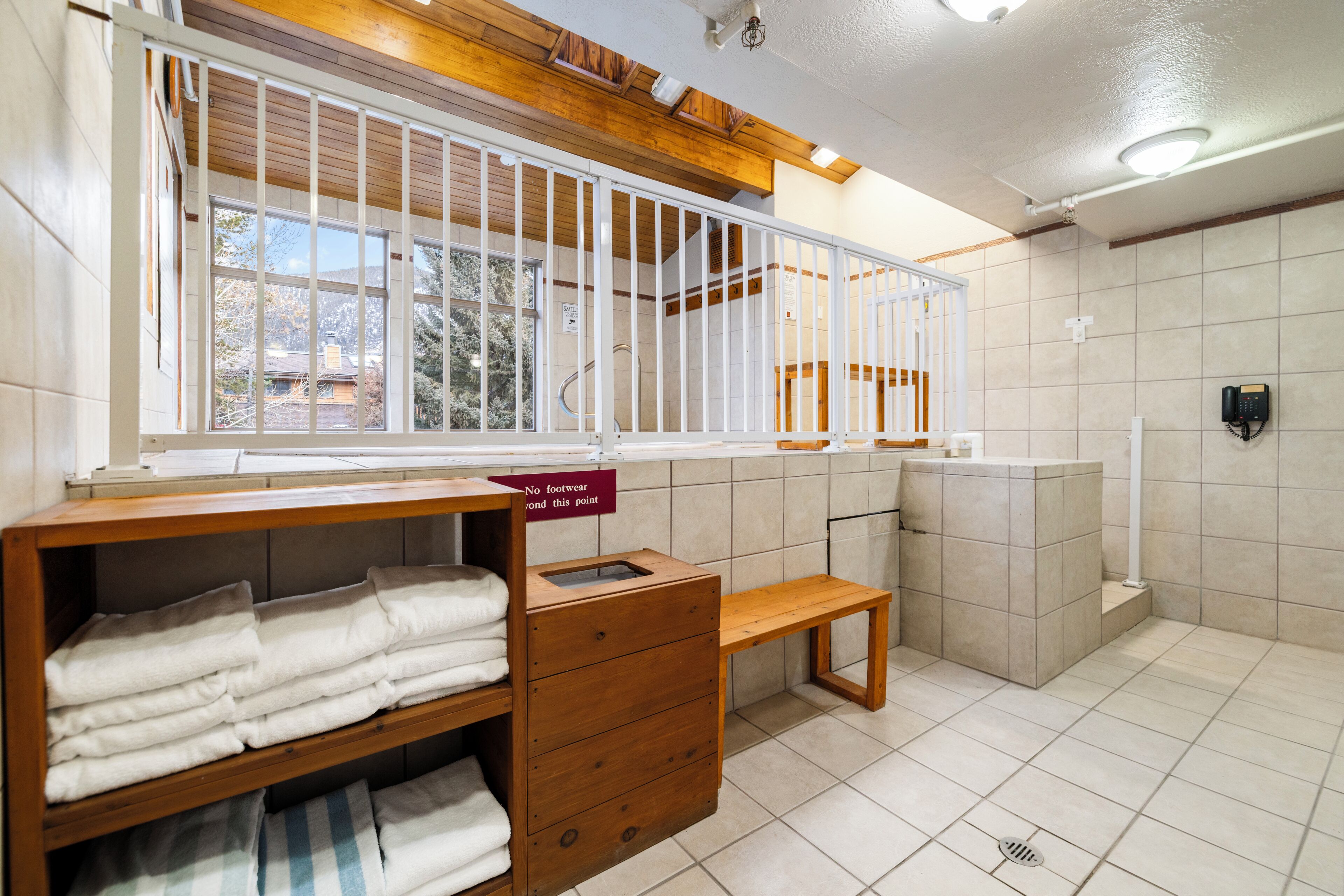 A hotel bathroom with a wooden bench, a towel rack, and a sign indicating no footwear beyond a certain point.