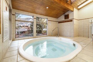A hot tub in a room with large windows, wooden ceiling, and tiled walls.