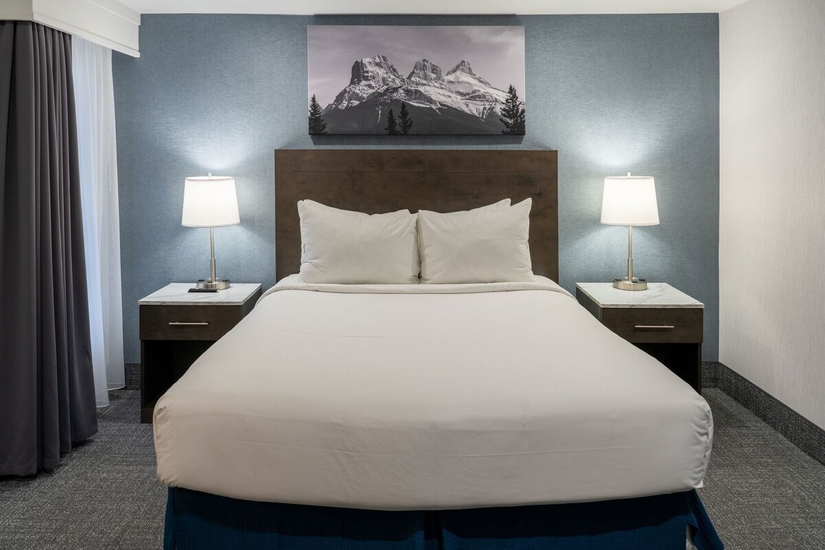 A hotel room with a large bed, two bedside tables with lamps, a headboard, a painting on the wall, and a window with curtains.