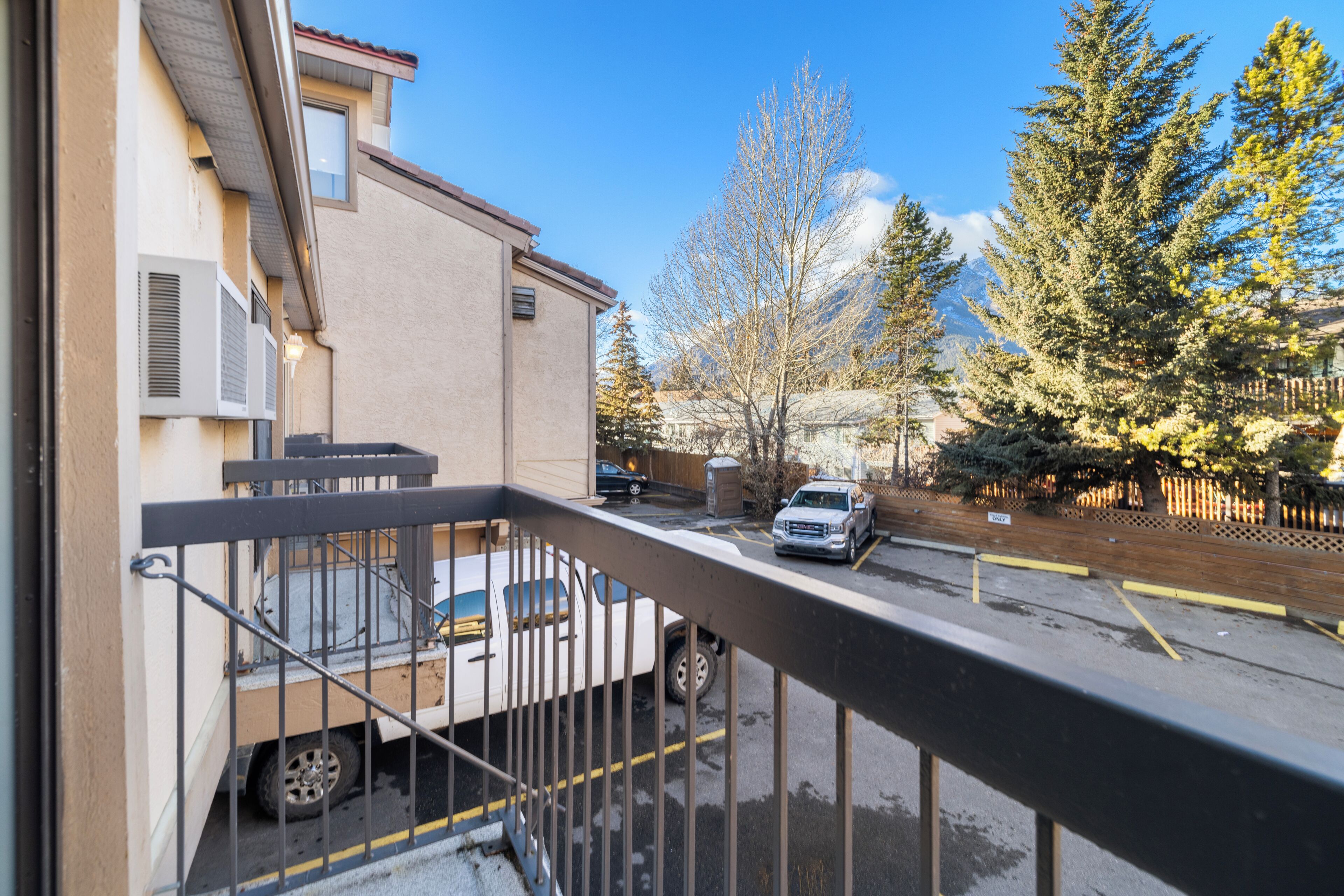A residential street with a parked truck and a car, a building with a balcony, and a row of evergreen trees.