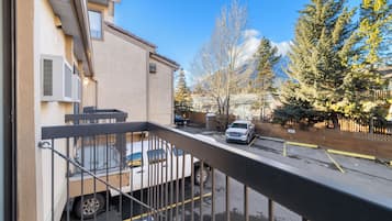 A residential street with a parked truck and a car, a building with a balcony, and a row of evergreen trees.