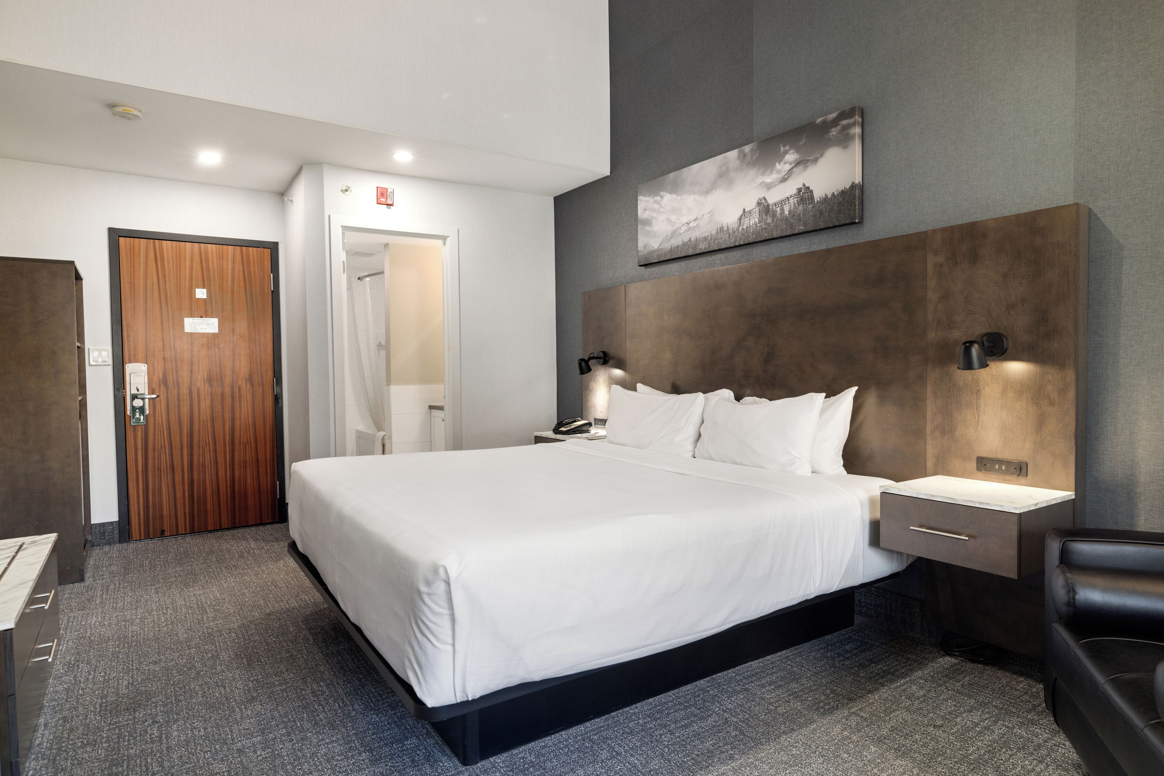 A hotel room with a large bed, a bedside table, a desk, a chair, a wooden door, a shower area, and a painting on the wall.