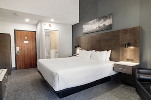 Habitación de hotel con una cama grande, una mesita de noche, un escritorio, una silla, una puerta de madera, zona de ducha y un cuadro en la pared.
