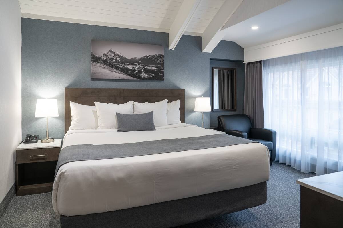 Habitación de hotel con una cama grande, dos mesitas de noche con lámparas, una silla, un escritorio, una ventana con cortinas y un cuadro colgado en la pared.
