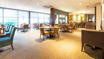 A spacious dining area with multiple tables and chairs, large windows, and a patterned carpet.