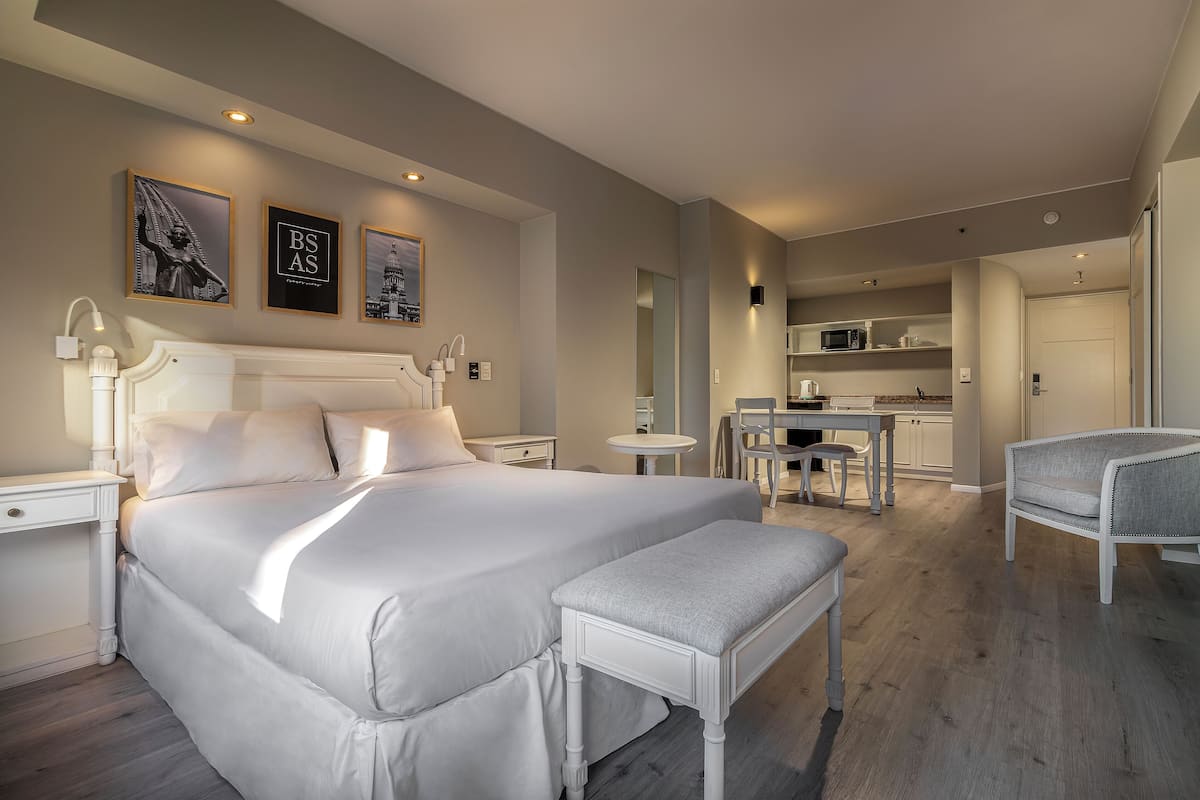 A modern hotel room with a large bed, a small table, a chair, and a kitchenette in the background.