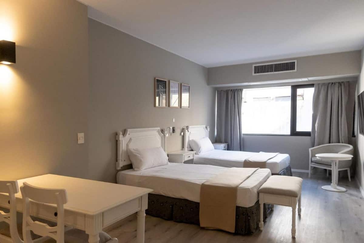 A hotel room with two beds, a dining table, chairs, a small round table, and a window with curtains.