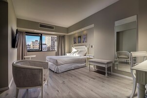 Quarto de hotel moderno com uma cama grande, uma escrivaninha, uma cadeira e vista para a cidade.