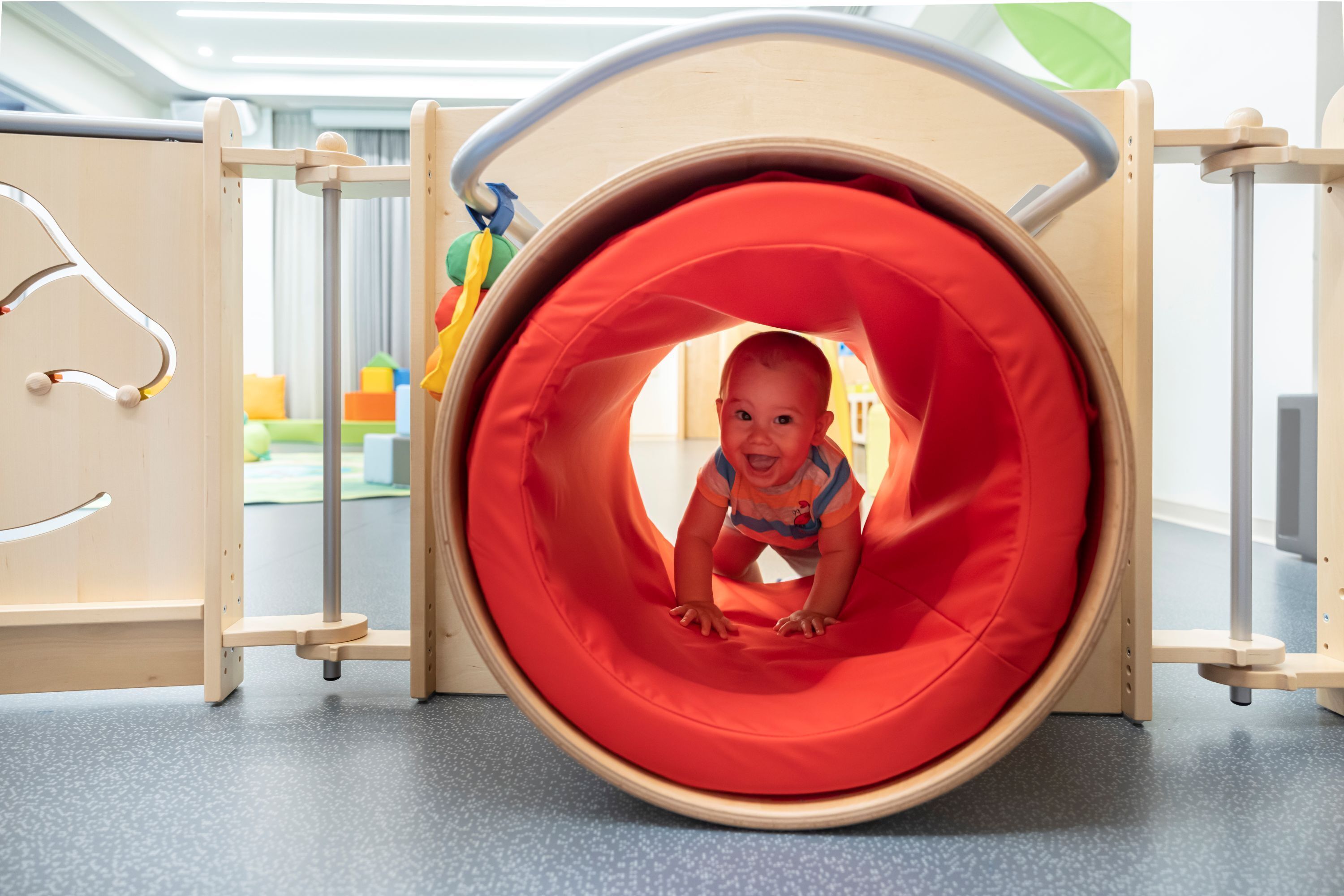 A child in a play tunnel with a red interior, surrounded by wooden play structures.