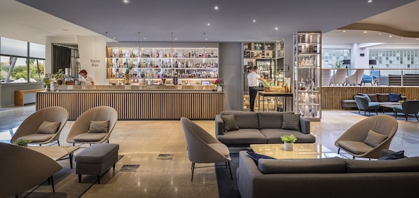 A modern bar area with a variety of liquor bottles, a seating area with sofas and armchairs, and a large window providing a view of the outdoors. - Valamar Diamant Hotel (Porec)