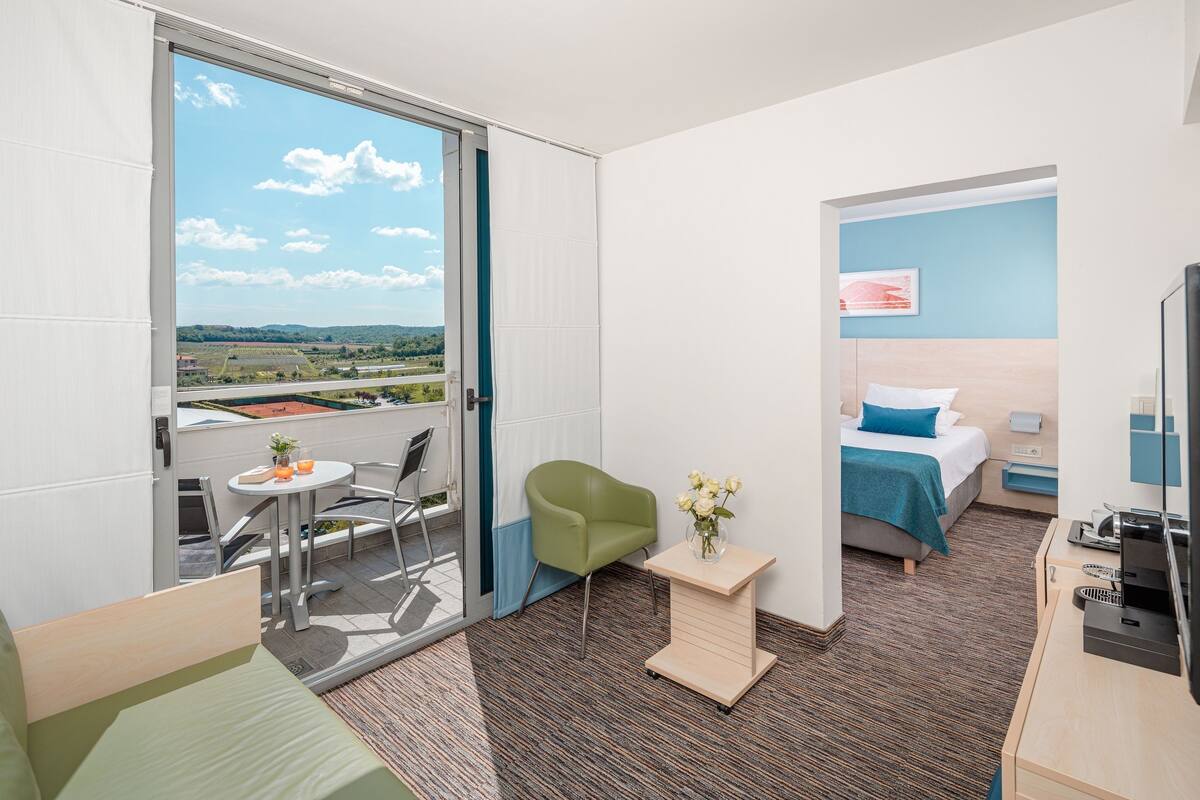 Una habitación de hotel moderna con balcón, una cama, un escritorio y una pequeña mesa con un florero.