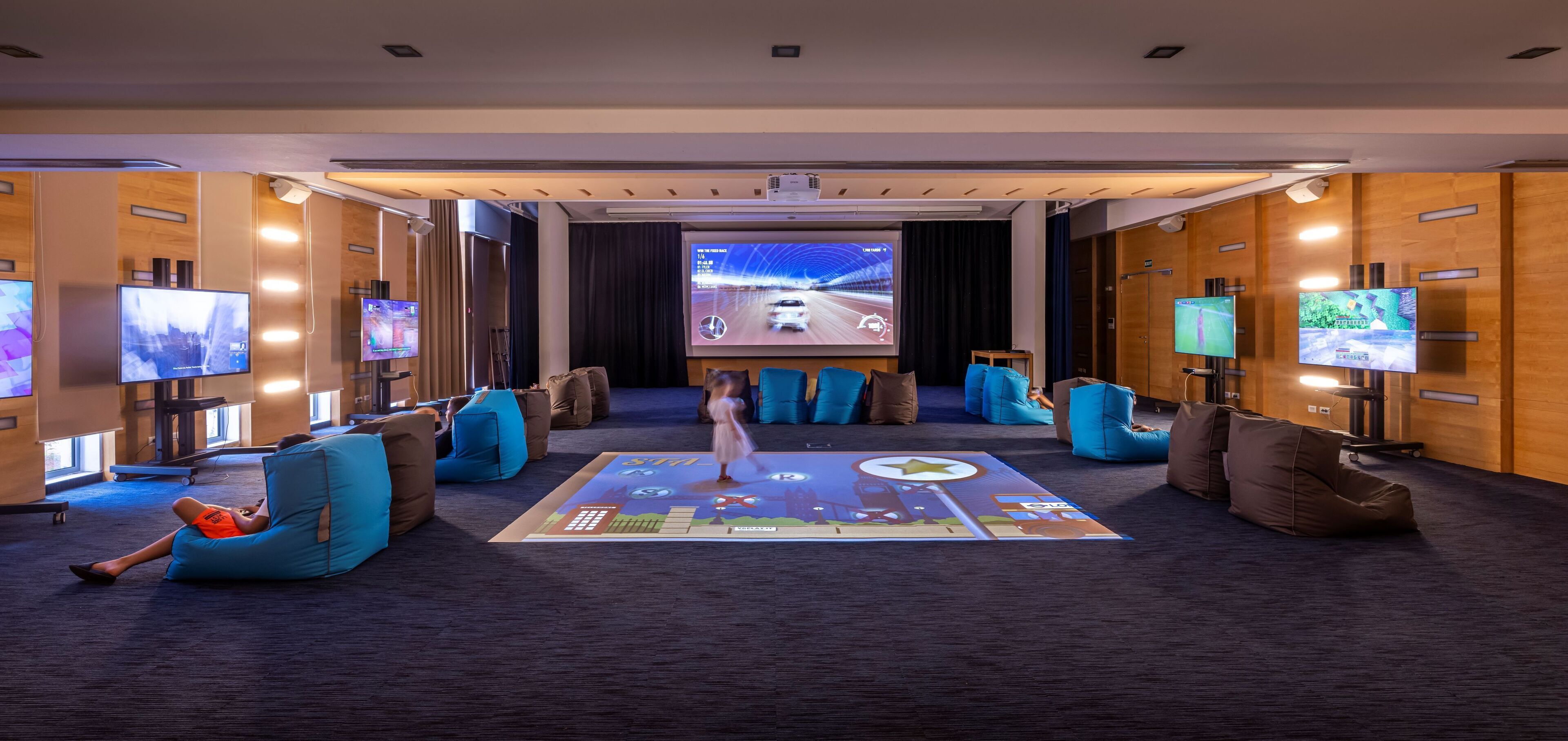 A modern hotel room with a large screen, multiple seating areas, and a carpeted floor.