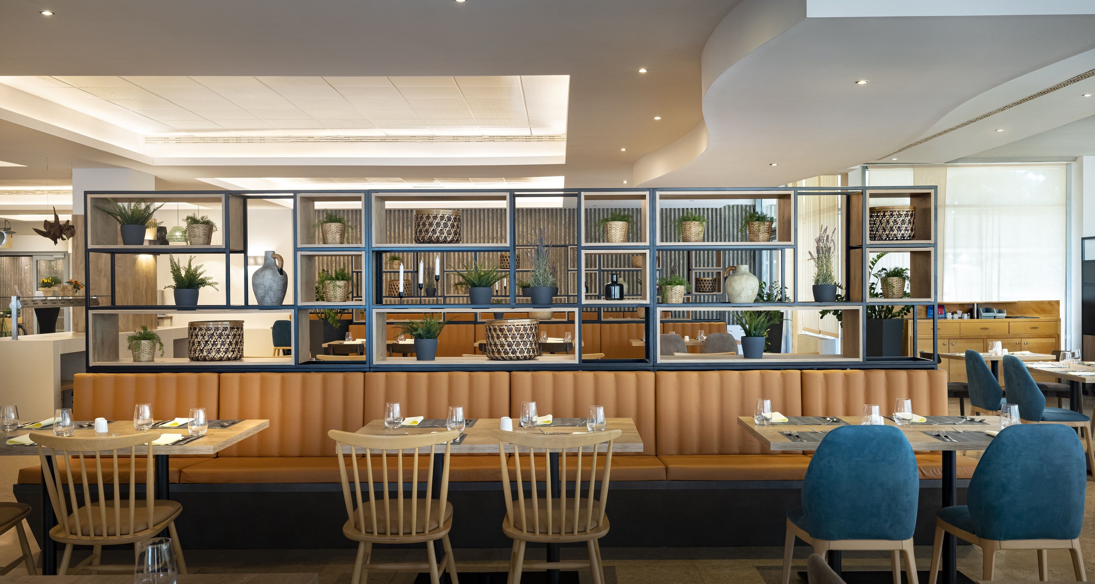 A restaurant with wooden booths and tables, a high ceiling with recessed lighting, and shelves with plants and decorative items.