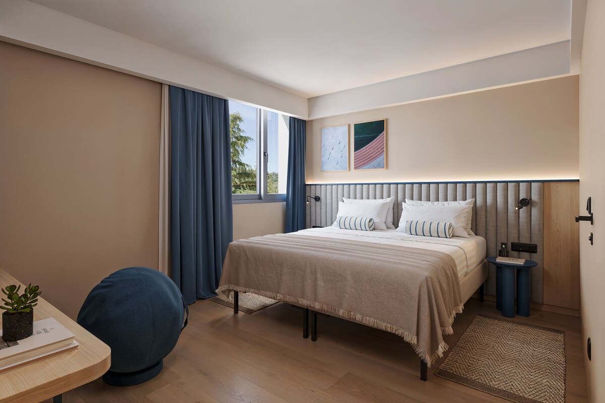 A modern hotel room with a large bed, a desk with a chair, a small table with a plant, and a window with curtains.