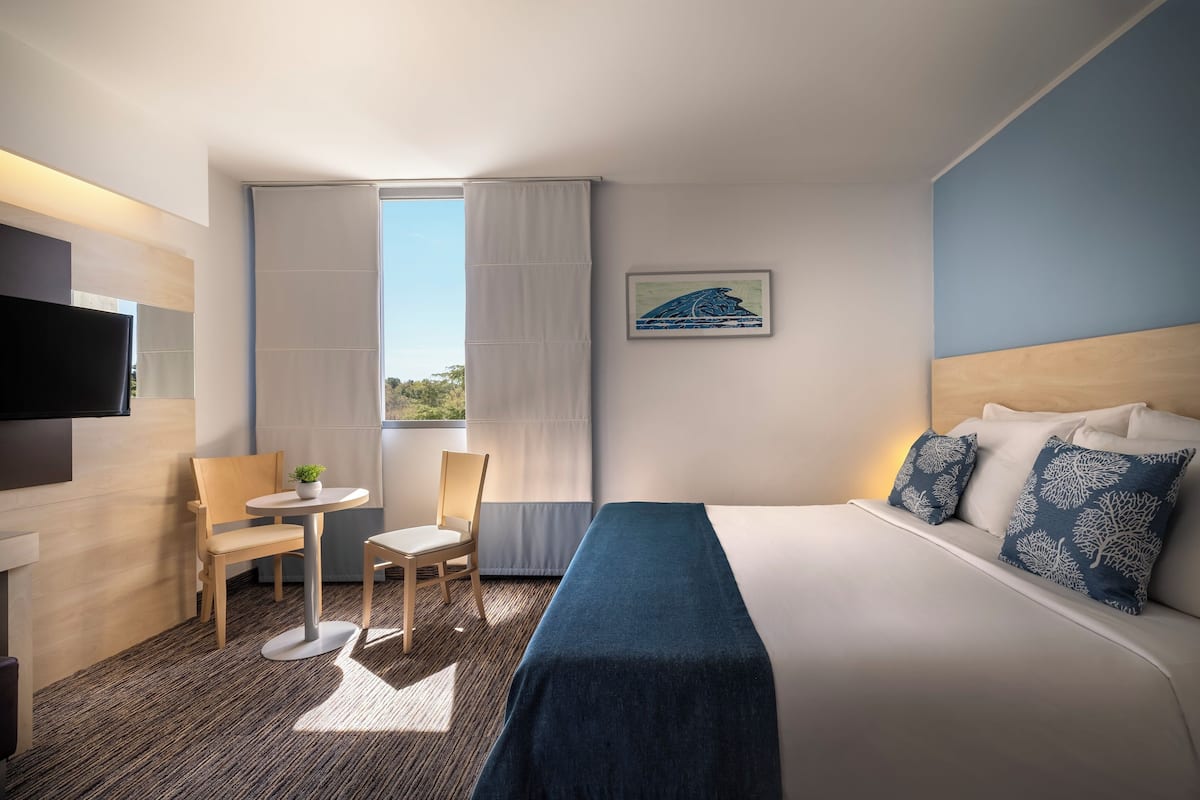 Una habitación de hotel con cama, escritorio, silla, televisor y ventana con cortinas.