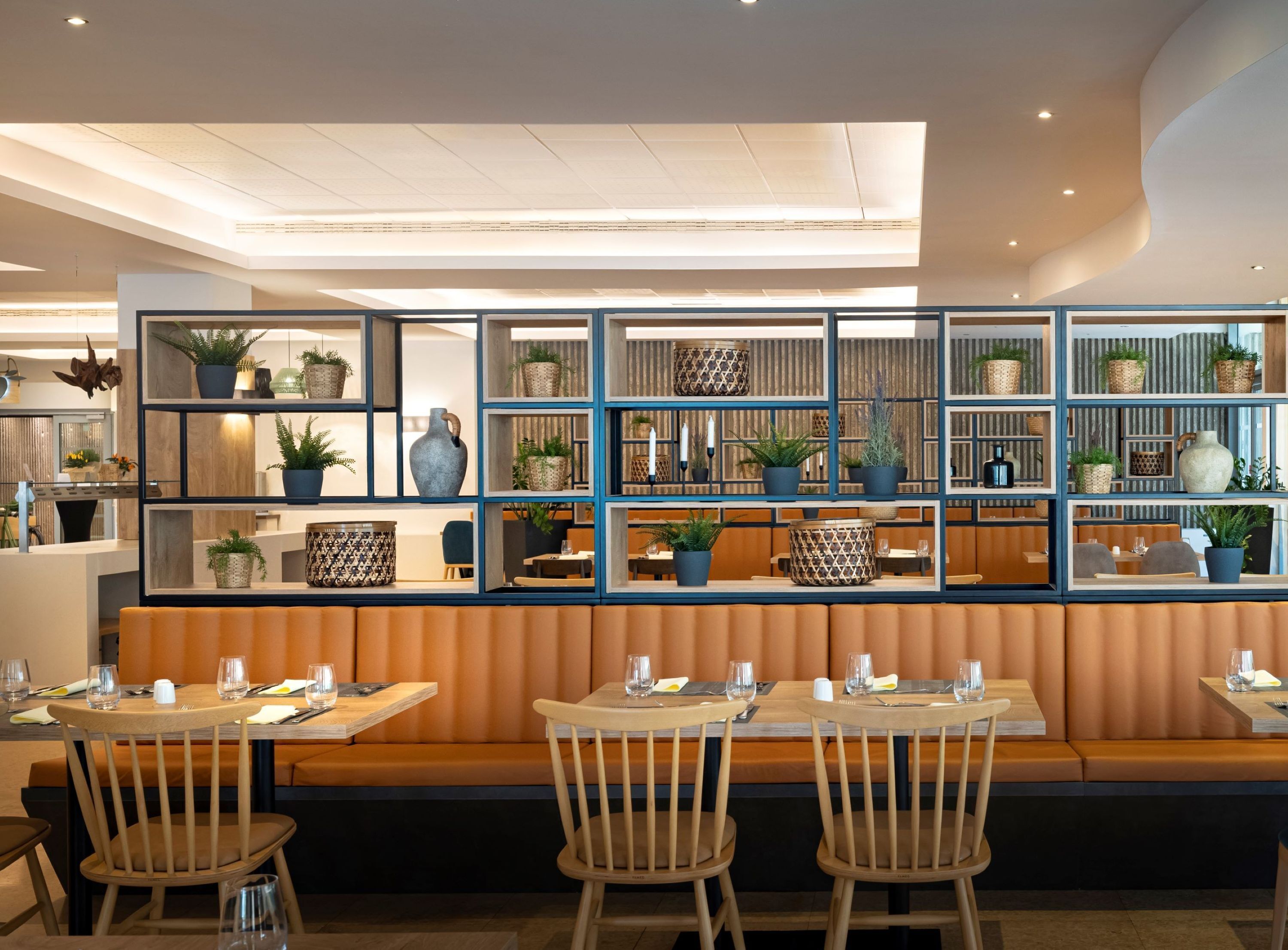 A modern restaurant interior with wooden booths, tables, and a wall of shelves displaying potted plants and decorative items.